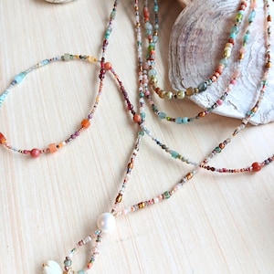 Peut inclure: Plusieurs colliers de perles et un bracelet aux couleurs variées, notamment bleu, orange et rose. Les colliers présentent diverses perles, dont des perles et de petits charms dorés. Les bijoux sont disposés sur une surface en bois clair avec des coquillages.