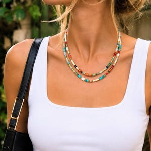 Puede incluir: Un collar de capas que presenta dos hileras de pequeñas cuentas de vidrio multicolor en tonos turquesa, rojo, naranja, blanco y verde, acentuadas con espaciadores de oro metálico. El collar se lleva con pendientes de aro dorados y una camiseta sin mangas blanca.
