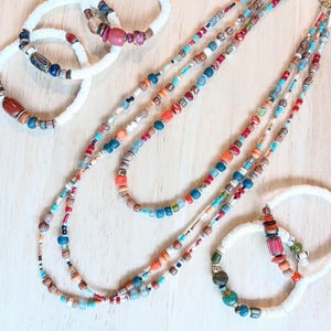 May include: A selection of colourful beaded necklaces and bracelets. The necklaces have multiple strands of small, vibrant beads in various colours, including red, blue, orange, and green. The bracelets are white with colourful accent beads. The jewellery is displayed on a light wooden surface.