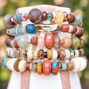 May include: A multi-strand bracelet featuring a diverse collection of beads in various shapes, sizes, and textures. The beads include smooth, matte, and speckled finishes in colors such as blue, orange, red, brown, white, and green, with a central gold-toned metal bar.