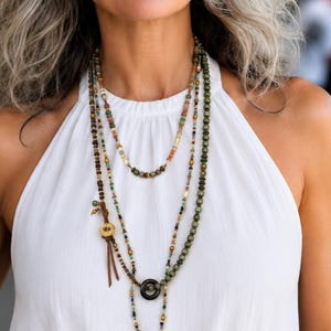 May include: A multi-strand beaded necklace featuring various earth-toned beads in shades of green, brown, and gold. The design includes a small circular charm with a leather tassel, a central black ring, and a decorative bronze-toned metal pendant.