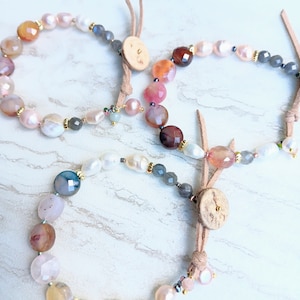 Può includere: Tre braccialetti con perline di pietra naturale, tra cui agata, perle e pietre grigie. Ogni braccialetto ha un cordoncino in pelle con una chiusura a bottone in legno.