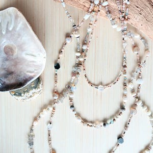 May include: Several layered necklaces with various beads, including pearls, and gemstones. The necklaces are in shades of white, cream, and pastel colors. A large seashell is in the upper left corner, and a piece of driftwood is in the background.