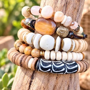 May include: A stack of five bracelets featuring a variety of beads including smooth pale pink stones, a large amber-colored oval bead, a metallic Buddha head charm, white disc-shaped beads, speckled brown jasper beads, and a black cylindrical bead with white etched patterns.