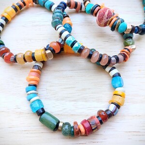 May include: Three colourful beaded bracelets. The bracelets feature a variety of beads in shades of orange, yellow, blue, green, and black. The beads are of different shapes and sizes, creating a unique and textured appearance.