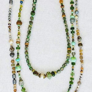 Puede incluir: Un collar largo de cuentas que presenta una variedad de cuentas de vidrio, piedra y metal en tonos verdes, marrones, crema y azules. El hilo incluye formas cilíndricas, esféricas y facetadas dispuestas en un patrón asimétrico.