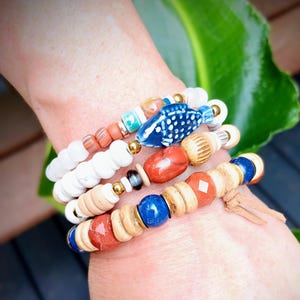 May include: A collection of beaded bracelets in various colors and textures. The bracelets feature a blue fish charm, wooden beads, red and white stones, and gold accents. The bracelets are stacked on a wrist.