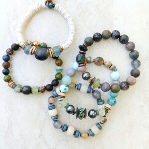 Puede incluir: Cuatro pulseras de cuentas de varios estilos. Una pulsera está hecha de cuerda blanca con cuentas. Las otras tres pulseras están hechas de cuentas de colores en tonos de azul, verde, marrón y gris. Las pulseras están sobre una superficie blanca.