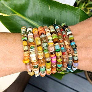 May include: A stack of ten multi-colored beaded bracelets featuring a variety of shapes, textures, and materials including wood, glass, and ceramic beads in shades of yellow, orange, green, blue, pink, brown, and white arranged in rows.