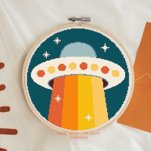 May include: A cross-stitch pattern of a UFO with a rainbow beam of light, on an 8-inch hoop. The pattern is on 18 count Aida cloth.  The UFO is white with orange, yellow, and red details. The background is dark blue with white stars.  on 8" hoop - 18 count @varidita