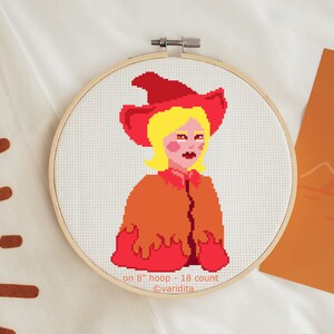 May include: Cross stitch pattern of a person with blonde hair wearing a red and orange witch costume. The pattern is on an 8-inch hoop and is 18 count. The text on the bottom reads "on 8" hoop - 18 count @varidita".