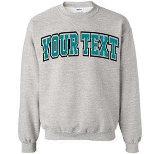 Puede incluir: Una sudadera gris claro con el texto "YOUR TEXT" en letras de bloque turquesa con contorno negro en el pecho. La sudadera tiene cuello redondo y mangas largas.