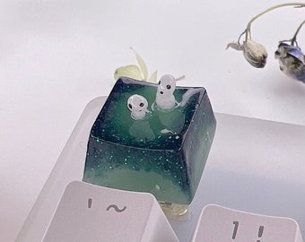 Puede incluir: Un tapón de tecla de resina verde y blanca con dos pequeñas figuras blancas en su interior. El tapón de tecla está sobre un teclado blanco.