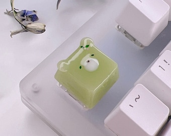 May include: A green and white resin keycap shaped like a bear with a white face and black eyes. The keycap is sitting on a white keyboard.