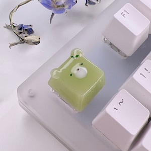 May include: A green and white resin keycap shaped like a bear with a white face and black eyes. The keycap is sitting on a white keyboard.