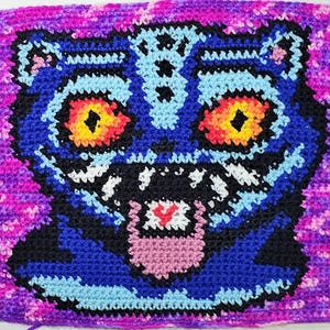 Peut inclure: Une pièce de crochet faite à la main représentant un visage de tigre stylisé. Le visage du tigre est principalement bleu, avec des accents noirs, des dents blanches et des yeux jaune et orange vifs. Le fond est un mélange de violet et de rose.