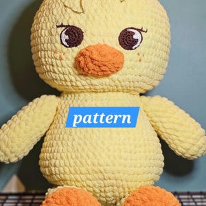 May include: A handmade, plush yellow chick with an orange beak and feet, and brown eyes. The chick has small wings and is sitting. The word "pattern" is written on a blue rectangle on the chick's body. The chick is made of a soft, textured material.