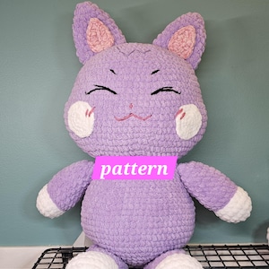 Könnte beinhalten: Ein handgefertigtes, plüschiges, lavendelfarbenes, katzenartiges Stofftier mit weißen Akzenten auf den Pfoten und Wangen. Die Ohren sind rosa gefüttert und das Gesicht hat gestickte Details. Das Wort "pattern" steht auf der Vorderseite.