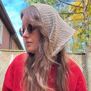 May include: A beige crocheted hood, worn over the head and shoulders, with a triangular shape at the back. The person is wearing a red sweatshirt and round sunglasses. The background includes trees and a blue sky.