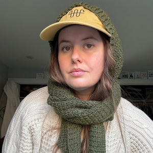 Könnte beinhalten: Eine Frau trägt einen grünen Strick Schal und eine beige Baseballkappe mit einem schwarzen gestickten Design. Die Kappe hat eine Kapuze, die über ihren Kopf gezogen ist. Sie trägt einen weißen Strickpullover.