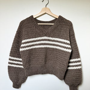 May include: A brown knitted sweater with white stripes on the sleeves and body. The sweater has a v-neck and is hanging on a wooden hanger.