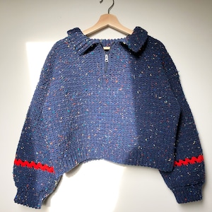 May include: A navy blue cropped sweater with a half-zip closure and red trim on the sleeves. The sweater has a speckled texture.