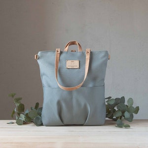 May include: A light blue canvas tote bag with leather straps and a small wooden tag. The bag has a large pocket on the front and a smaller pocket on the back.