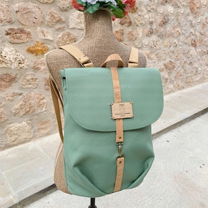 Op de afbeelding: Een saliegroene canvas rugzak met een kurkband en een houten label met de tekst "KM.LI CONSCIOUSLY HANDMADE SPAIN".