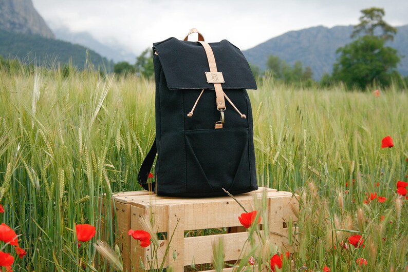 May include: A black canvas backpack with leather straps and a flap closure. The backpack is sitting on a wooden crate in a field of green grass and red poppies.