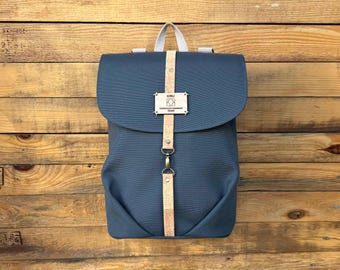 May include: A blue backpack with a flap closure and a vertical tan strap. The strap has a metal buckle and a small rectangular tag with text. The backpack has a white handle and is set against a wooden background.