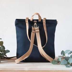 May include: A dark blue canvas backpack with brown leather straps and buckles. The backpack has a top handle and a flap closure.