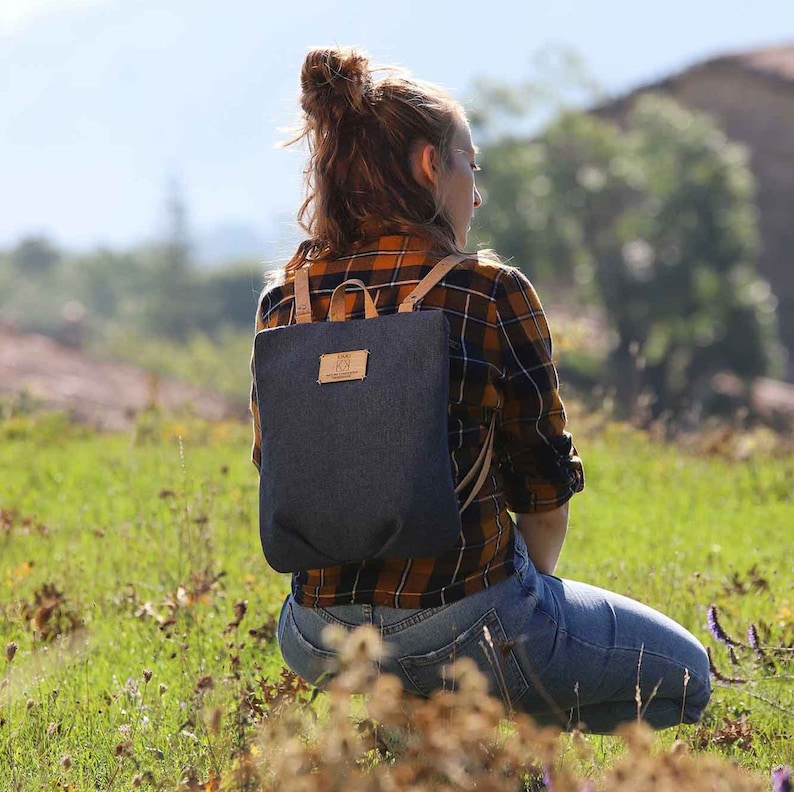 May include: A person wearing a blue and yellow checked shirt with a dark blue denim backpack. The backpack has a brown leather strap and a small brown leather patch with the text "EKO" on it.