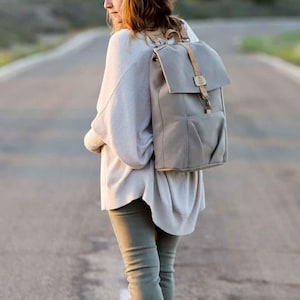 May include: A woman wearing a light gray sweater and olive green pants is walking down a road with a gray canvas backpack with brown leather straps and buckles.