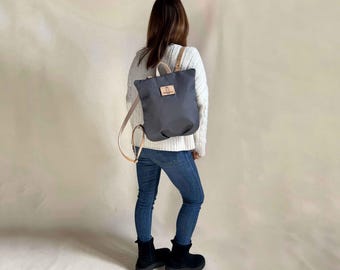 Puede incluir: Una mochila de lona gris con una solapa de cuero marrón y un pequeño parche rectangular de cuero. La mochila la lleva puesta una persona con el pelo largo y castaño.