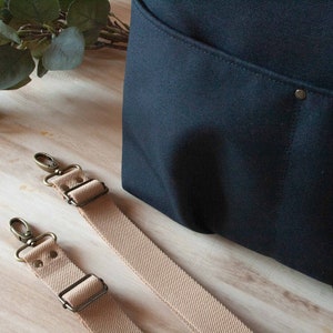 May include: Two beige canvas straps with brass hardware and metal clasps. The straps are lying on a wooden surface next to a black canvas bag.
