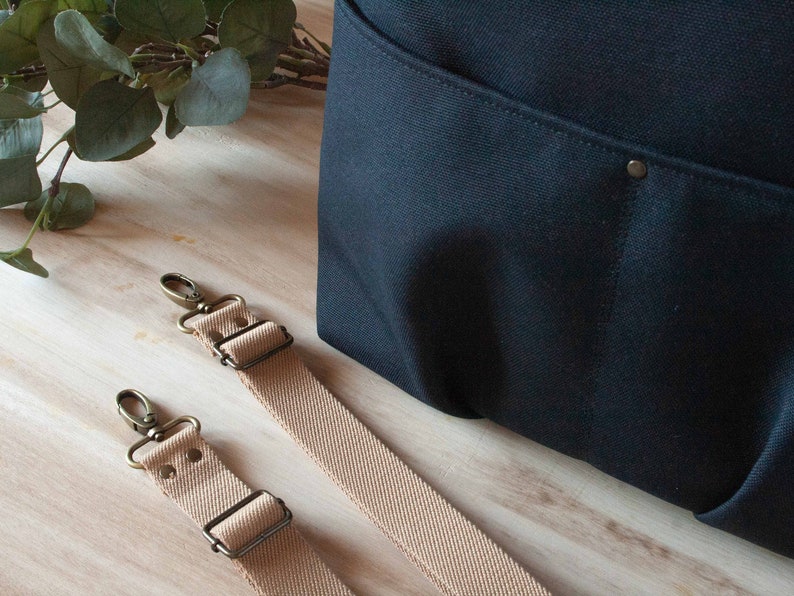 May include: Two beige canvas straps with brass hardware and metal clasps. The straps are lying on a wooden surface next to a black canvas bag.