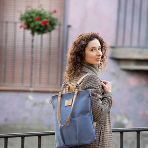 May include: A woman wearing a plaid jacket and blue jeans is carrying a blue canvas backpack with brown leather straps. The backpack has a large front pocket and a small leather patch with text on it.