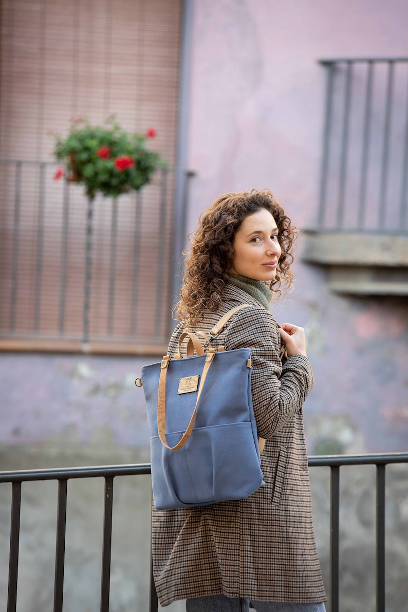 May include: A woman wearing a plaid jacket and blue jeans is carrying a blue canvas backpack with brown leather straps. The backpack has a large front pocket and a small leather patch with text on it.