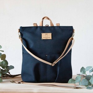 May include: A dark blue canvas backpack with a tan leather strap and a small wooden tag that says "KOALA, CONSCIOUSLY HANDMADE, DENMARK".