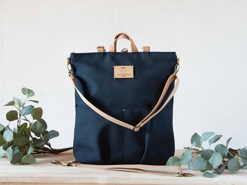 May include: A dark blue canvas backpack with a tan leather strap and a small wooden tag that says "KOALA, CONSCIOUSLY HANDMADE, DENMARK".