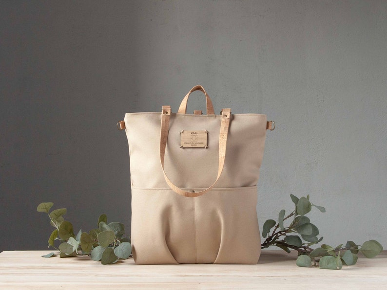 May include: A beige canvas tote bag with leather straps and a cork patch that says "KIMI - Crafted with Passion - Berlin".