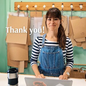 Op de afbeelding: Een vrouw draagt een blauwe denim overalljurk met een wit en zwart gestreept lang mouw shirt. Ze staat voor een muur met een houten rek en bruine papieren patronen. De tekst "Thank you!" is in witte letters op de muur geschreven.