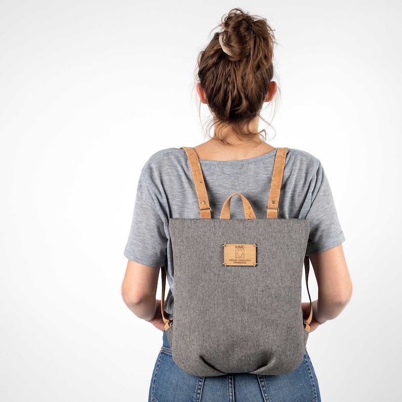 May include: A grey canvas backpack with brown leather straps and a cork label that reads "K&B Natural Cork Products Handmade".