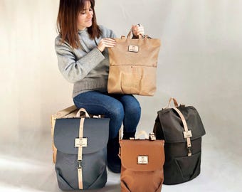 May include: A woman holding a tan backpack with a top handle and a small rectangular label. Three other backpacks in different colors are displayed: dark gray, brown, and olive green. Each has a flap closure and light-colored straps.