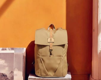 Könnte beinhalten: Ein beigefarbener Rucksack mit Überschlag, einem mittigen hellen Riemen und einem Metallhakenverschluss. Der Rucksack verfügt über einen rechteckigen Aufnäher mit dem Text "KXX" auf der Klappe und eine Vordertasche mit schrägen Seitennähten.