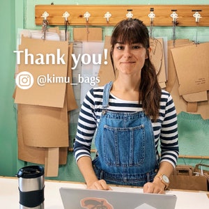 May include: A woman wearing a blue denim overall dress with a white and black striped shirt is standing in front of a wall with brown paper patterns. She is smiling and looking at the camera. The text "Thank you! @kimu_bags" is on the wall.
