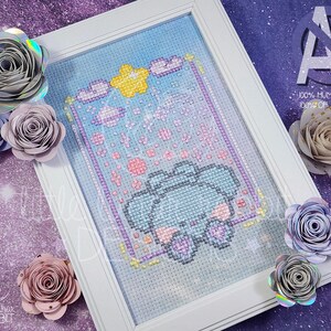 May include: A framed cross-stitch artwork featuring a light blue character with rounded ears, surrounded by a yellow star, pink clouds, and small sparkles on a grid background, accented by several paper flowers in shades of gray, pink, and purple.