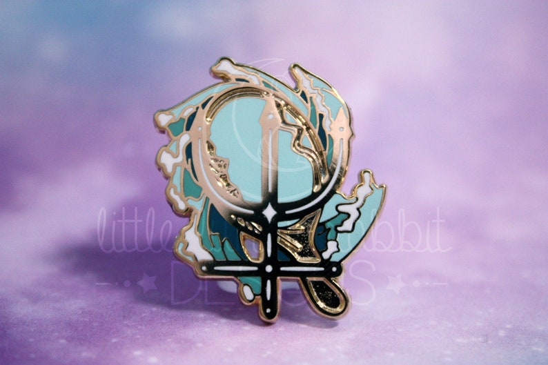 Peut inclure: &Eacute;pinglette en &eacute;mail dor&eacute; repr&eacute;sentant un trident stylis&eacute; avec une lune croissante et des vagues bleues, vertes et blanches.