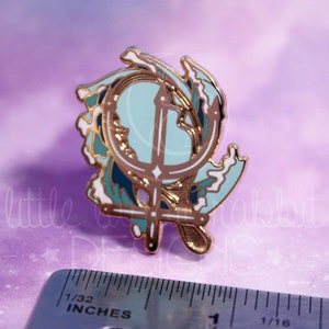 Peut inclure: &Eacute;pinglette en &eacute;mail dor&eacute; avec un design d'ancre stylis&eacute; entour&eacute; de vagues bleues et blanches. L'ancre est un trident stylis&eacute; avec une croix au sommet. L'&eacute;pingle mesure environ 2,5 cm de diam&egrave;tre.
