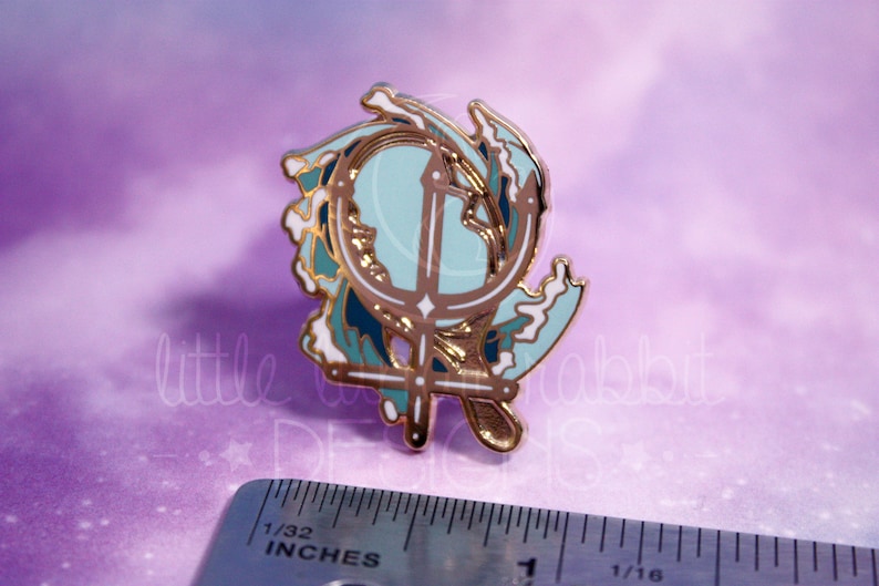 Peut inclure: &Eacute;pinglette en &eacute;mail dor&eacute; avec un design d'ancre stylis&eacute; entour&eacute; de vagues bleues et blanches. L'ancre est un trident stylis&eacute; avec une croix au sommet. L'&eacute;pingle mesure environ 2,5 cm de diam&egrave;tre.